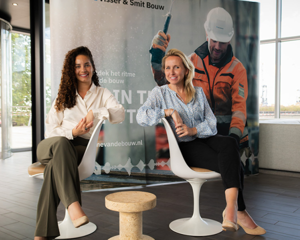 Djisri en Anita op zitten op twee stoelen in een lichte ruimte. Op de achtergrond een man met een oranje jas aan en helm op